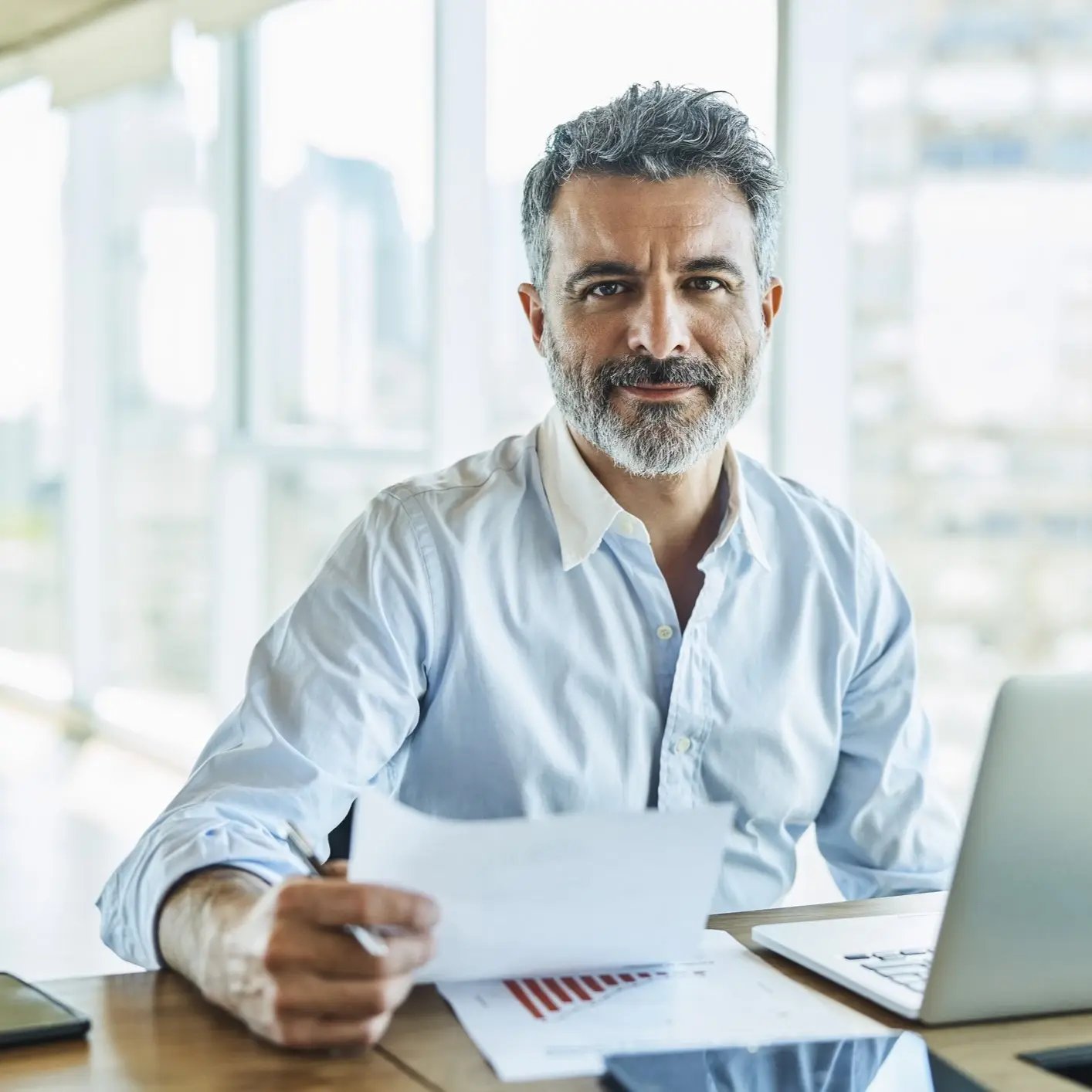 Confident businessman reviewing a loan document at his desk with a laptop and smartphone.