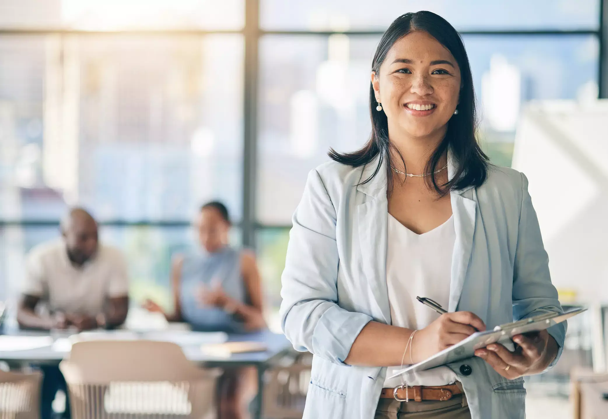 Smiling Asian businesswoman leads meeting, holding clipboard. Diverse team members visible in background.