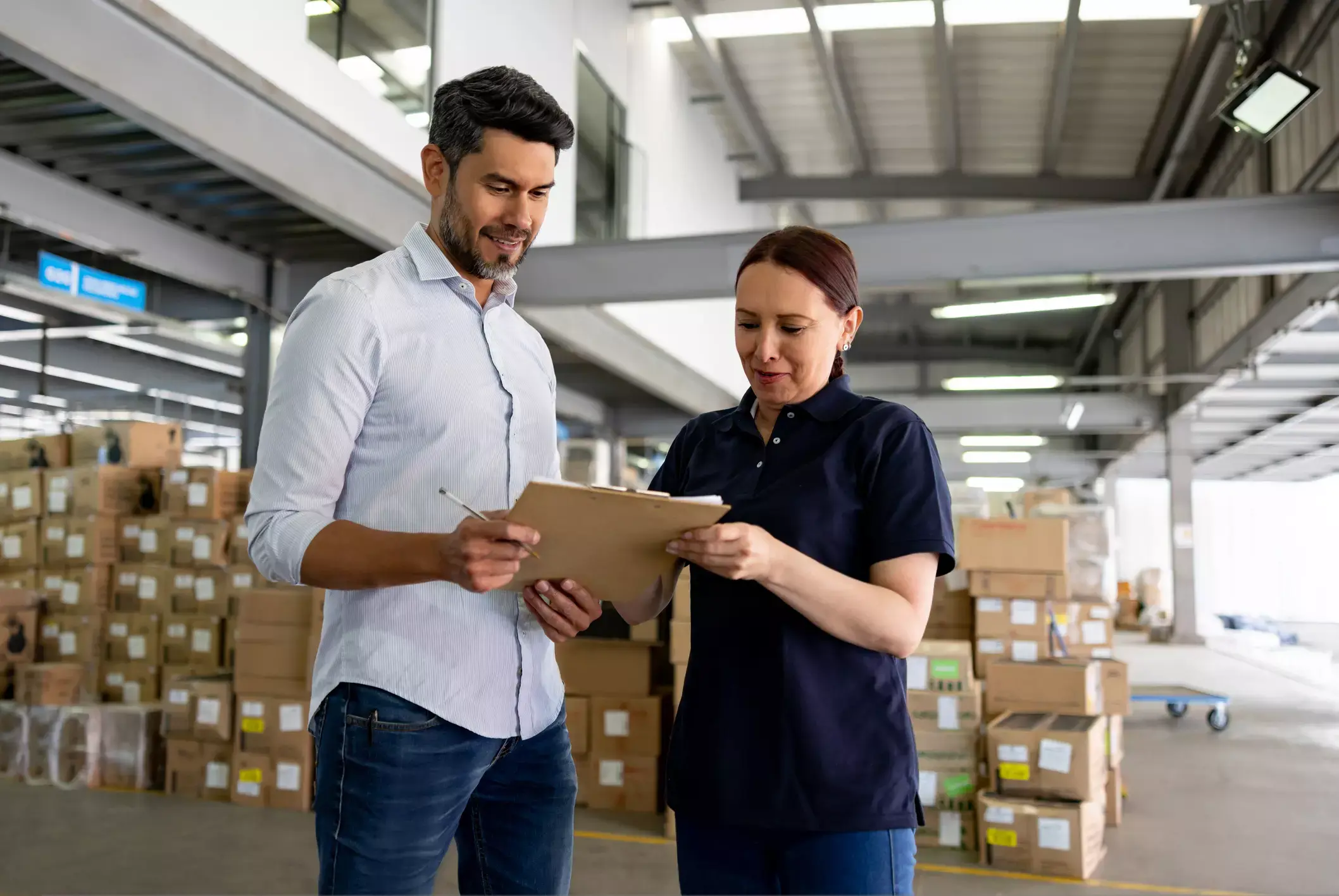 Two professionals reviewing clipboard in warehouse filled with stacked boxes, collaborating on inventory management.