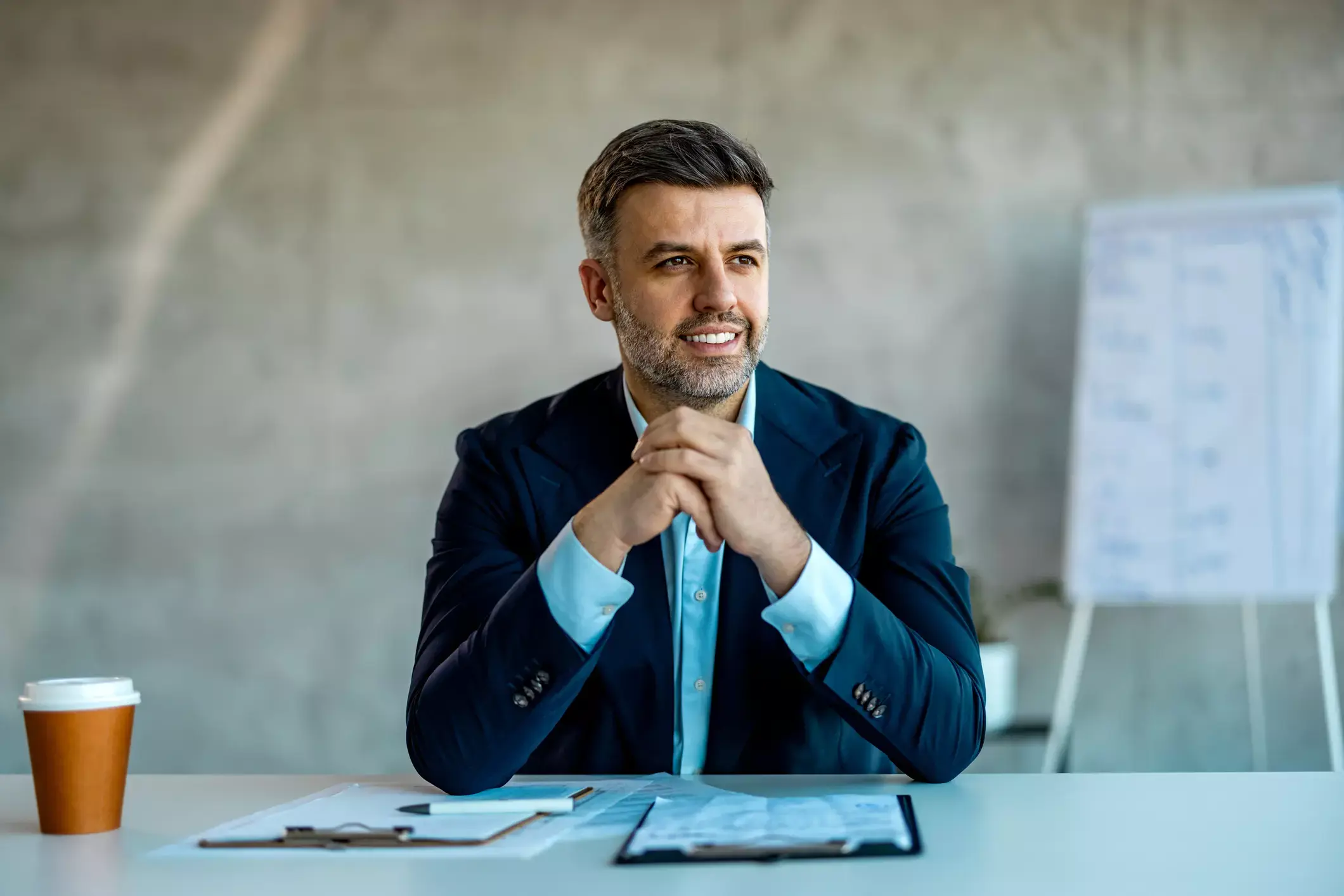 Executive in navy suit ponders business decision, documents and coffee on desk, whiteboard in background