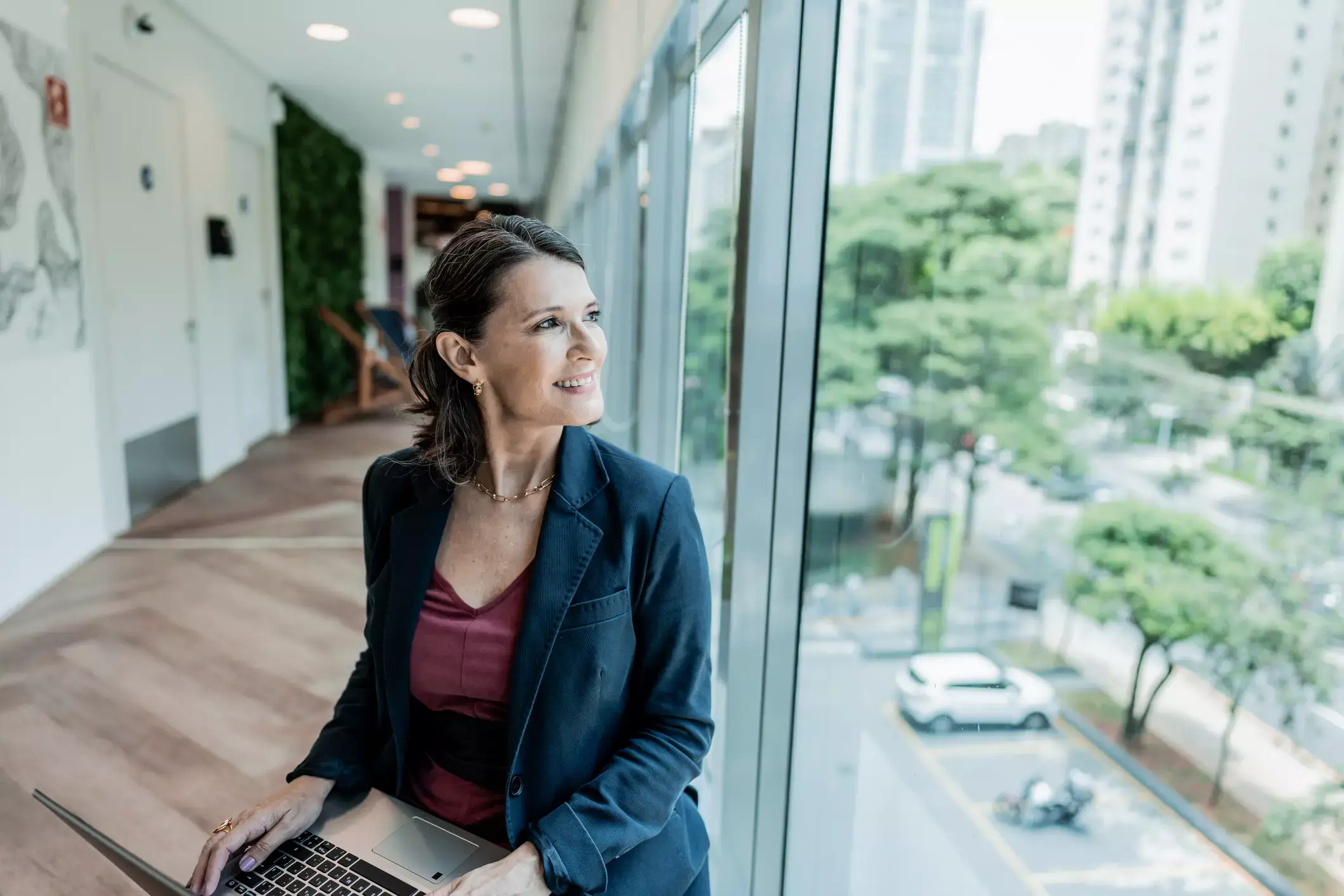 Confident professional woman with laptop gazes out office window at urban landscape, smiling optimistically.